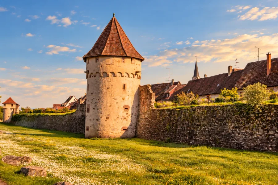 Medieval town walls of Bergheim in Alsace, with historic defensive towers and red-tiled roofs beneath a blue sky.