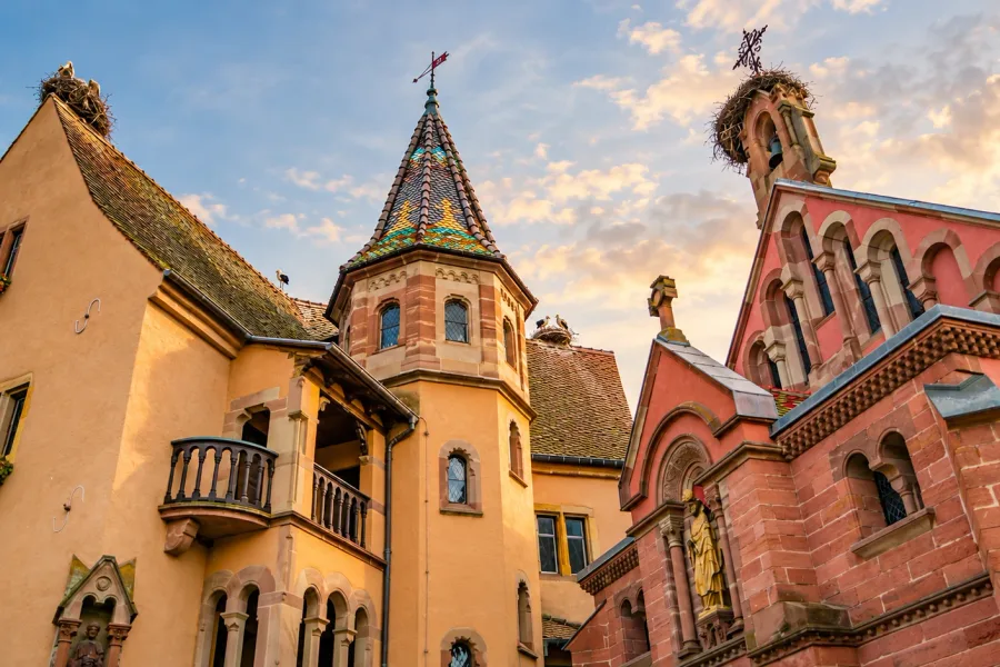 Historic architecture in Eguisheim, Alsace, with a tower featuring colorful roof tiles, a pink sandstone chapel and stork nests on the rooftops.
