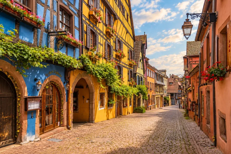Cobblestone street in Riquewihr, Alsace, lined with blue and yellow half-timbered houses, flower-filled façades and traditional shop signs.